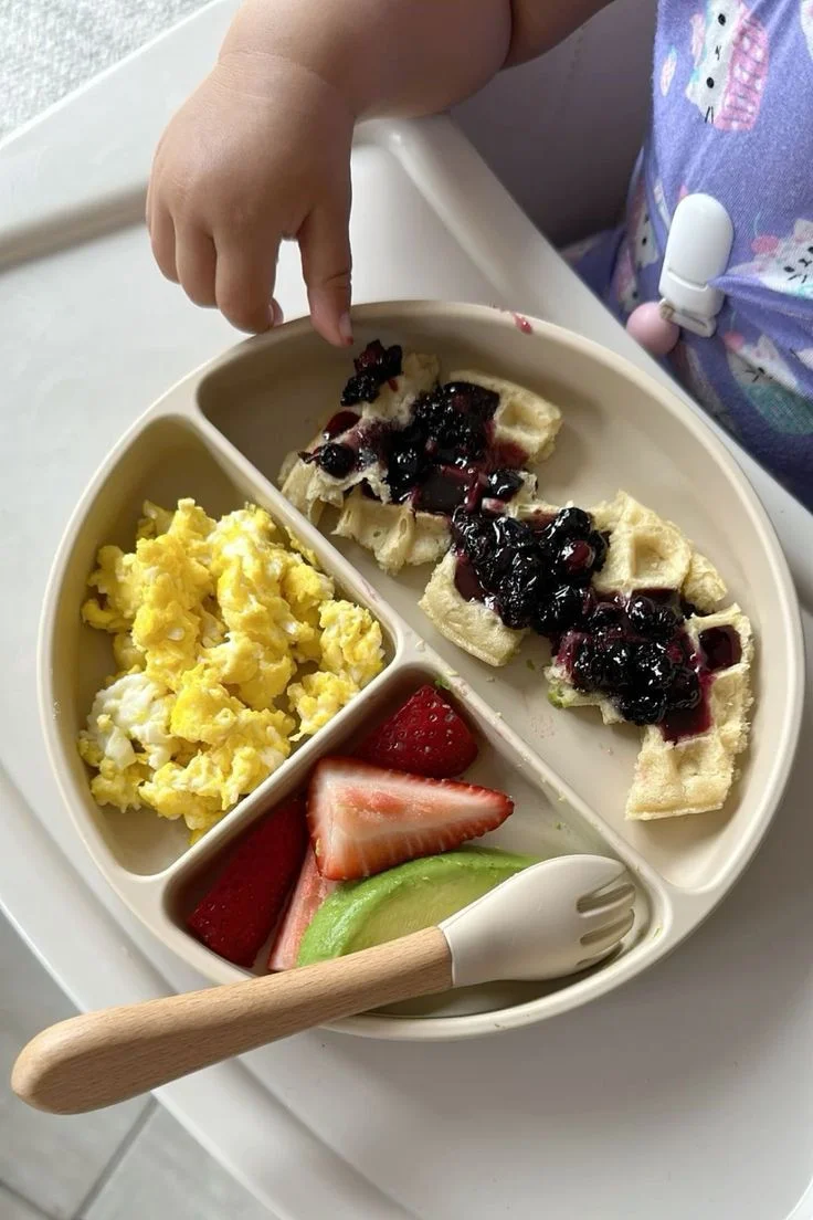 Peque tomando waffles con arándanos junto a huevo revuelto, fresas y aguacate.
