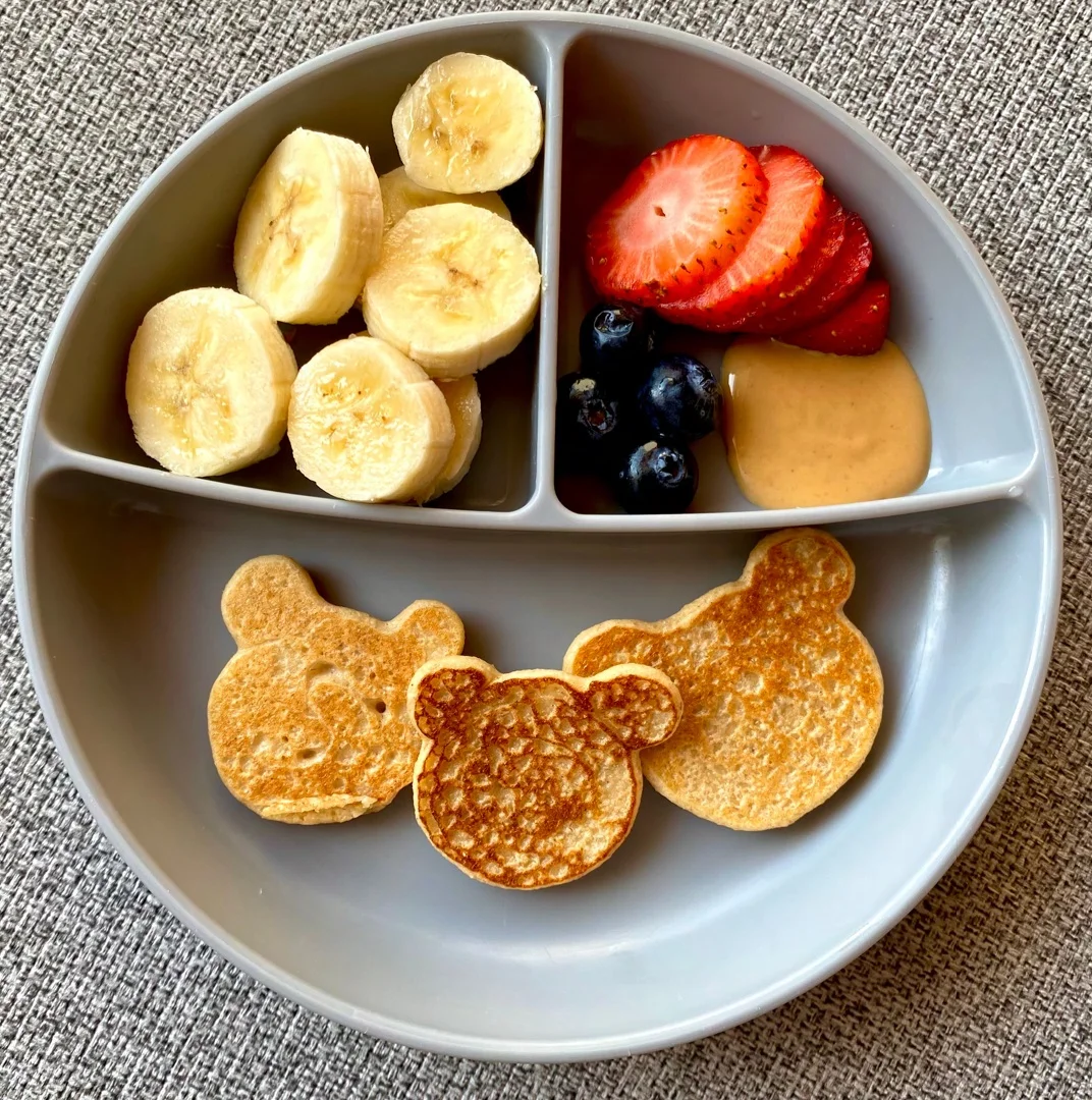Plato infantil con pancakes en forma de osito, banano, fresas, arándanos y crema de maní.